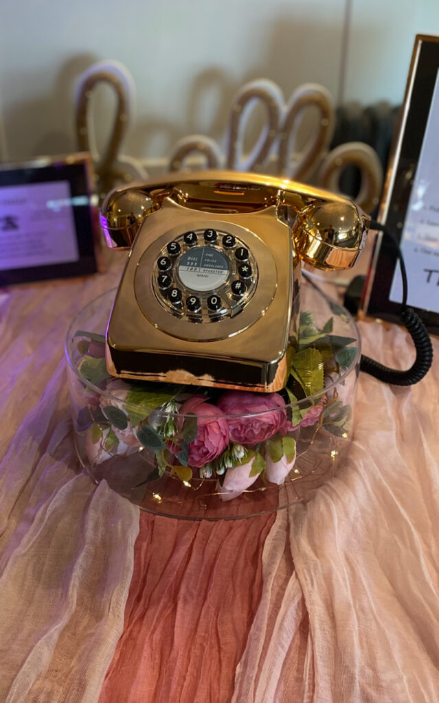Flower filled wedding stand upon pink drapes supporting a metallic rose gold retro telephone audio guest book in Maidstone wedding venue.